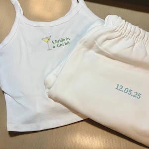May include: White pajama set comprising a vest top and trousers. The vest top features a cocktail graphic and the text "A Bride in a tini bit" in green. The trousers display the date "12.05.25" in blue.