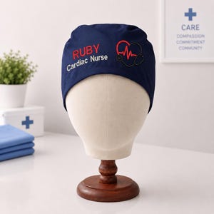 Puede incluir: Un gorro quirúrgico azul marino con el texto bordado RUBY Cardiac Nurse en rojo y blanco, junto a un gráfico de corazón rojo con el contorno de un estetoscopio en negro. El gorro se muestra sobre un soporte de cabeza de maniquí.