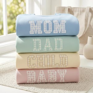 May include: A stack of four pastel-colored sweatshirts. Each sweatshirt has a different word embroidered on it in a gingham pattern: MOM, DAD, CHILD, and BABY. The sweatshirts are stacked on a neutral-colored rug.