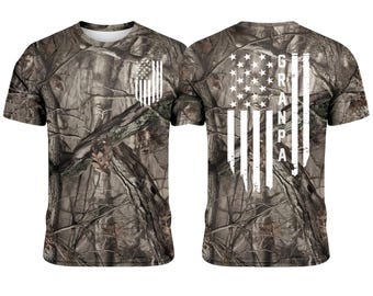 Könnte beinhalten: T-Shirt mit Camouflage-Muster und weißem amerikanischem Flaggen-Design auf Vorder- und Rückseite. Auf der Rückseite des Shirts steht das Wort "GRANDPA" in weißen Großbuchstaben. Das Shirt hat einen Rundhalsausschnitt und kurze Ärmel.