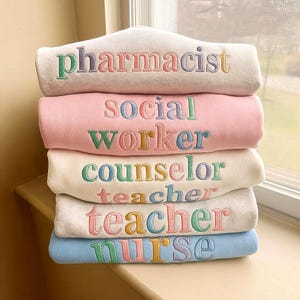 Puede incluir: Una pila de sudaderas de colores pastel bordadas con títulos de trabajo. La sudadera superior es de color beige claro con "pharmacist" en hilo colorido. Debajo hay sudaderas rosas, crema, gris claro y azules, cada una con una profesión diferente bordada.