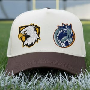 May include: A baseball cap with a cream-colored crown and a brown bill. The cap features two embroidered patches: an eagle head on a yellow and black shield and a blue and gray dragon in a circle. The cap is sitting on green grass.