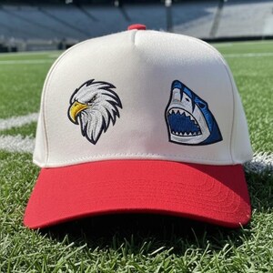 May include: A baseball cap with a white crown and a red brim. The cap features embroidered patches of an eagle and a shark. The cap has a red button on top and is sitting on green grass.