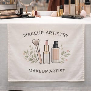 May include: A beige table runner with the words "MAKEUP ARTISTRY" and "MAKEUP ARTIST" printed in black. The runner features illustrations of a makeup brush, lipstick, and foundation bottle, with floral accents.
