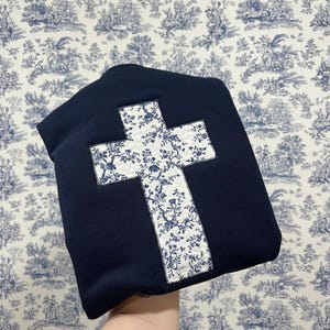 Op de afbeelding: Een marineblauw sweatshirt met een grote kruisapplicatie. Het kruis is gemaakt van witte stof met een blauw bloemenpatroon. Het sweatshirt wordt getoond tegen een blauw en wit toile behang.