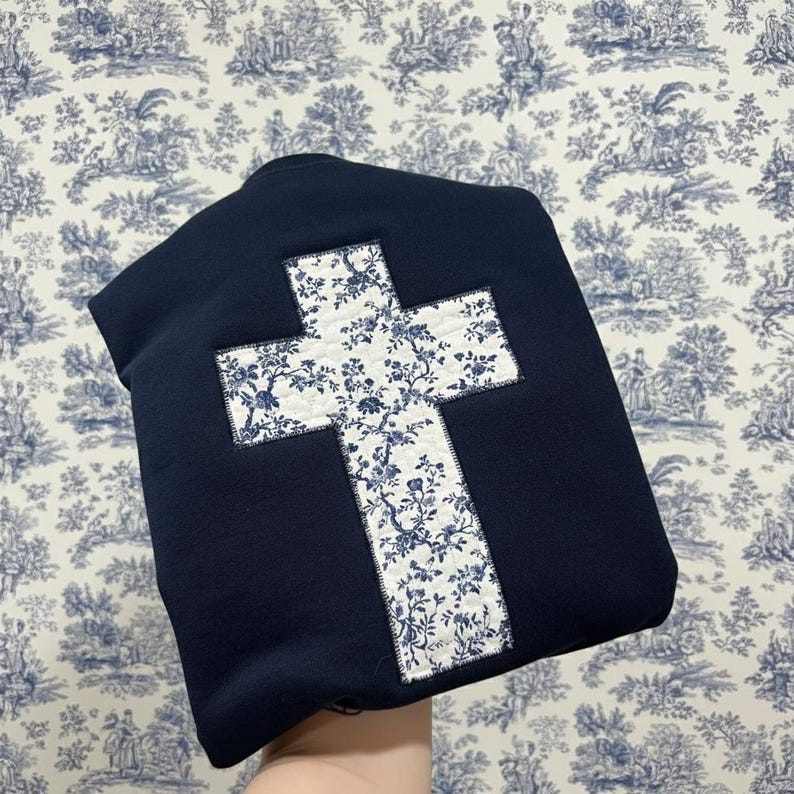 Op de afbeelding: Een marineblauw sweatshirt met een grote kruisapplicatie. Het kruis is gemaakt van witte stof met een blauw bloemenpatroon. Het sweatshirt wordt getoond tegen een blauw en wit toile behang.