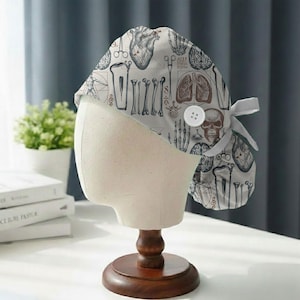 Puede incluir: Un gorro quirúrgico con un patrón de ilustraciones anatómicas en gris y marrón sobre un fondo blanco. El diseño incluye corazones, pulmones, calaveras y huesos. El gorro se muestra en una cabeza de maniquí con un soporte de madera.