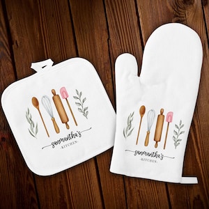 Puede incluir: Conjunto de agarraderas de cocina blancas con un diseño de acuarela de utensilios de cocina, que incluye un rodillo de madera, un batidor, una cuchara y una espátula. El texto "samantha's kitchen" está impreso en ambas piezas.