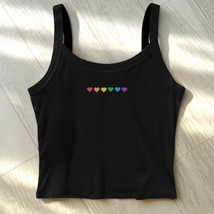Puede incluir: Camiseta sin mangas negra con una fila de seis corazones de colores del arcoíris bordados en el pecho. La camiseta tiene tirantes finos y un escote redondo. La prenda está hecha de un material suave y elástico.