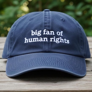 May include: A navy blue baseball cap featuring the embroidered phrase "big fan of human rights" in white. The cap has a curved peak and an adjustable strap. Made from a soft, durable fabric, it's a casual accessory.