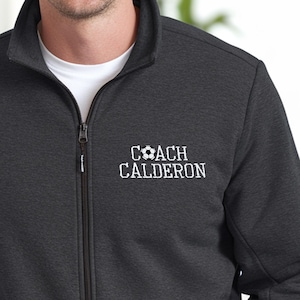May include: A dark gray fleece jacket with a zipper closure. The jacket has a white embroidered design that says "COACH CALDERON" with a soccer ball graphic.
