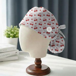 May include: A white and red patterned scrub cap with a cherry and heart design. The cap has a button closure and a tie in the back. The cap is displayed on a mannequin head.