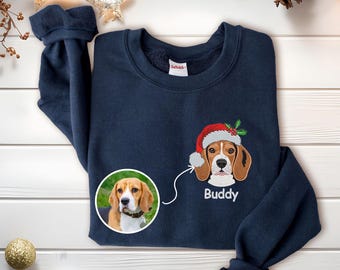 May include: A navy blue sweatshirt with a personalized embroidered image of a dog wearing a Santa hat. The dog's name, "Buddy", is embroidered below the image. A smaller image of the dog is in a circle on the left side of the sweatshirt.