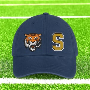 May include: Navy blue baseball cap with a tiger head logo and the letter "S" in gold with a white outline. The tiger logo is orange, black, and white. The cap is on a green field with white lines.
