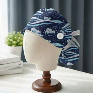May include: A navy blue scrub cap with a wave pattern in shades of blue and white. The cap has a white button and a matching tie. The cap is displayed on a mannequin head with a wooden stand.