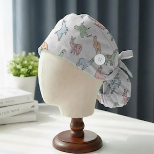 May include: A light gray scrub cap featuring a repeating pattern of colorful llamas. The cap has a button and a tie closure. The llamas are in various colors, including blue, green, pink, and orange.