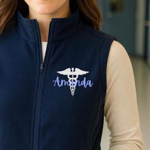 Op de afbeelding: Marineblau fleece vest met een volledige ritssluiting. De vest toont de naam "Amanda" in een scriptlettertype, samen met een wit caduceussymbool. Dit is een medisch of gezondheidsgerelateerd kledingstuk.
