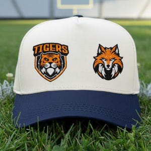 May include: A white and navy blue baseball cap with two embroidered patches. One patch features a tiger head with the word "TIGERS" above it. The other patch is a fox head. The cap is sitting on green grass.