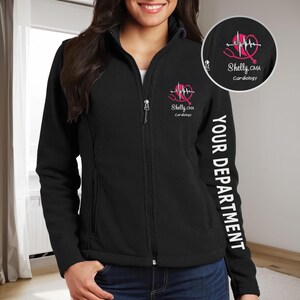 Puede incluir: Chaqueta de forro polar negra con una cremallera blanca y un corazón rosa con un estetoscopio y un gráfico de línea de ECG en el pecho izquierdo. El texto "Shelly, CMA Cardiology" está debajo del gráfico. La manga derecha tiene el texto "YOUR DEPARTMENT" impreso verticalmente.