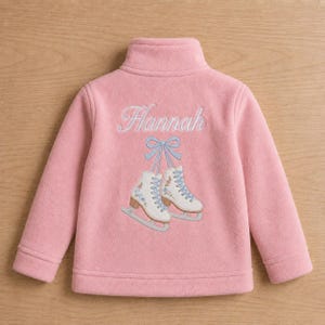 May include: A light pink fleece jacket featuring a high collar and long sleeves. The back displays an embroidered design of two white ice skates tied together with a light blue ribbon, positioned below the name Hannah in cursive script.