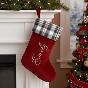 Può includere: Una calza di Natale rossa con polsino a quadri e il nome "Emily" ricamato in oro. La calza è appesa a un caminetto bianco decorato con verde e luci, vicino a un albero di Natale decorato.