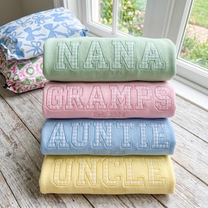 Può includere: Quattro cuscini cilindrici color pastello impilati su una superficie di legno. Ogni cuscino è ricamato con un nome diverso: "NANA", "GRAMPS", "AUNTIE" e "UNCLE", con "Est. 2026" sotto. Le lettere sono riempite con un motivo a quadretti vichy.