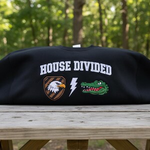 Könnte beinhalten: Schwarzes Sweatshirt mit dem weißen Text "HOUSE DIVIDED". Es zeigt einen gestickten Adler, einen Blitz und ein grünes Alligator. Das Sweatshirt wird auf einem hölzernen Picknicktisch mit verschwommenem Hintergrund von Bäumen präsentiert.