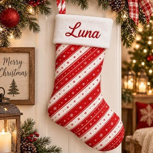 May include: A Christmas stocking featuring a red and white diagonal striped pattern with small white snowflake accents. The stocking has a plush white cuff at the top with the name Luna embroidered in red script lettering and a red and white loop.