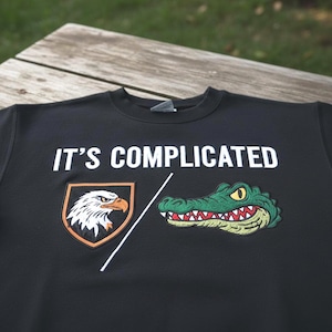 Puede incluir: Sudadera negra de cuello redondo con la frase "IT'S COMPLICATED" en letras blancas. Cuenta con un parche bordado de águila a la izquierda y un parche de cocodrilo verde a la derecha, separados por una línea diagonal blanca.