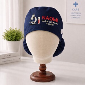 May include: A navy blue scrub cap featuring a white microscope icon, a red test tube icon, and the text "NAOMI Medical Laboratory Scientist" in red and white. The cap includes a white button on the side for securing a face mask.