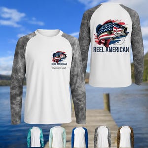 May include: White long-sleeve shirt with grey marbled sleeves. The front features a graphic of a fish with an American flag design and the text "REEL AMERICAN". The back of the shirt has a larger version of the fish graphic and the text "REEL AMERICAN".