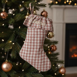 Puede incluir: Un calcetín navideño con un patrón de cuadros vichy en color rosa empolvado y blanco. El calcetín tiene un puño con volantes en la parte superior con el nombre Emma bordado en letras cursivas blancas. Incluye un lazo de tela para colgar.