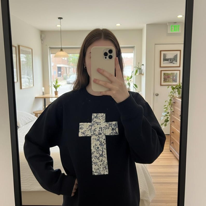 Op de afbeelding: Zwart sweatshirt met een groot kruisontwerp op de voorkant. Het kruis is gemaakt van witte stof met een blauw bloemenpatroon. De sweatshirt heeft lange mouwen en een ronde hals.