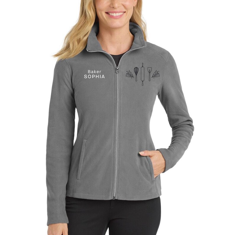 May include: A gray fleece zip-up jacket with a high collar. The jacket features the text "Baker SOPHIA" and a graphic of baking tools. The jacket has two front pockets and a silver zipper.