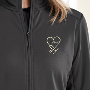 Può includere: Giacca nera con zip con un design a forma di cuore e stetoscopio dorati e il testo "Your Name" sul petto sinistro.