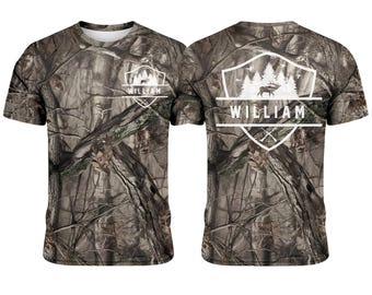 Könnte beinhalten: Ein Camouflage-T-Shirt mit einem weißen Emblem mit einem Hirsch, Bäumen und dem Namen "WILLIAM". Das Shirt hat einen Rundhalsausschnitt und kurze Ärmel. Auf der Rückseite des Shirts befindet sich ebenfalls das gleiche Emblem und der gleiche Name.