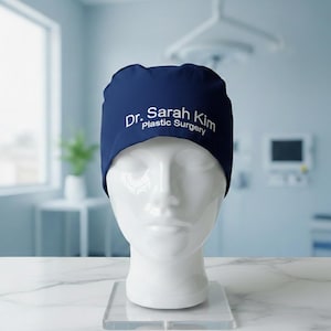 May include: A navy blue surgical cap with white text reading "Dr. Sarah Kim Plastic Surgery". The cap is displayed on a white mannequin head. The background is a blurred medical setting.