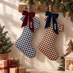 May include: Two Christmas stockings featuring a gingham check pattern, one in navy blue and one in brown. Each stocking has a ruffled cuff, a velvet bow, and a personalised name tag reading Olivia on the blue and Ethan on the brown stocking.