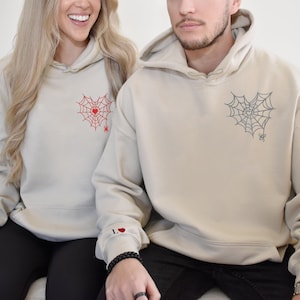 May include: Two beige hoodies with a heart-shaped spiderweb design. One hoodie has a red spiderweb and the other has a black spiderweb. The hoodie on the left has a red heart with the letter "L" on the sleeve.