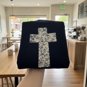 Op de afbeelding: Een marineblauwe tote bag met een grote kruisapplicatie. Het kruis is gemaakt van een bloemenstof met een witte achtergrond en blauw bloemenpatroon. De tas wordt voor een caf&eacute; gehouden.