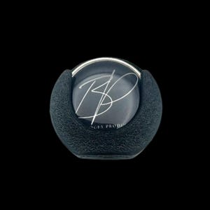 May include: A black, textured, round object with a silver rim and a clear, circular center. The center features a silver signature and the text "AGEY PRODUCTS". The object is set against a black background.