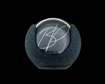 May include: A black, textured, round object with a silver rim and a clear, circular center. The center features a silver signature and the text "AGEY PRODUCTS". The object is set against a black background.