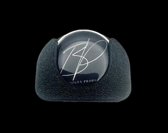 May include: A black, textured bottle stopper with a silver rim. The stopper features a clear, circular insert with the initials "BP" in a silver script. The text "OGEY PRODUC" is printed below the initials.