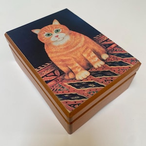 Puede incluir: Caja de madera con tapa con bisagras. La tapa presenta una colorida ilustración de un gato naranja con ojos verdes, sentado sobre una alfombra estampada sobre un fondo azul oscuro. La caja es de madera marrón claro.