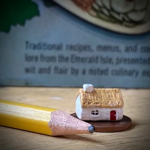 May include: A miniature white cottage with a thatched roof, a red door, and a small window. The cottage is sitting on a penny.