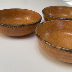 Puede incluir: Tres cuencos de cerámica marrón con superficie texturizada y borde oscuro. Los cuencos son redondos y tienen un aspecto moteado, lo que sugiere una estética artesanal o rústica. Los cuencos están vacíos y listos para usar.