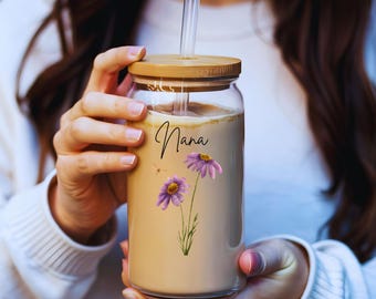 May include: A clear glass reusable tumbler with a bamboo lid and a straw. The tumbler has a floral design with purple flowers and the word "Nana" written on it.