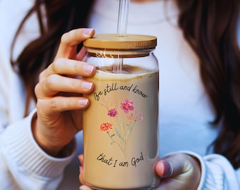 May include: A glass jar with a wooden lid and a straw filled with a brown liquid. The jar has a floral design and the text "Be still and know that I am God".