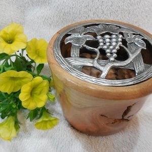 May include: Wooden bowl with a decorative metal grape design on the lid. The bowl is light brown with a darker rim. Yellow flowers and green leaves are on the left side of the bowl.