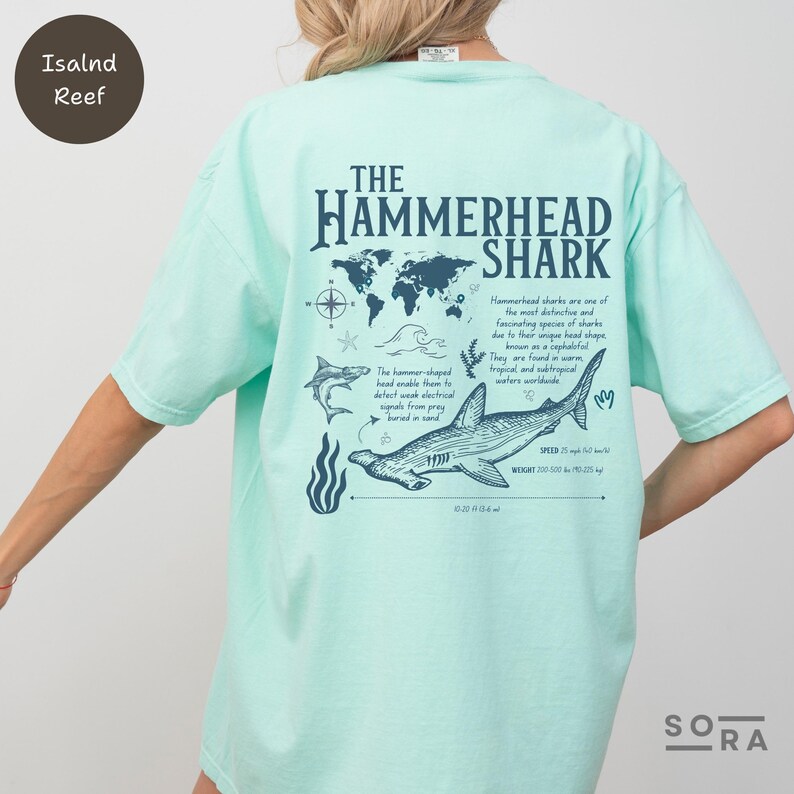 K&ouml;nnte beinhalten: Ein hellblaues T-Shirt mit einem Grafikdruck eines Hammerhais und einer Weltkarte. Der Text "The Hammerhead Shark" ist auf dem T-Shirt aufgedruckt. Der Text unter dem Hai lautet "Hammerhaie sind eine der markantesten und faszinierendsten Haiarten aufgrund ihrer einzigartigen Kopfform, die als Cephalofol bekannt ist. Sie kommen in warmen tropischen und subtropischen Gew&auml;ssern weltweit vor."